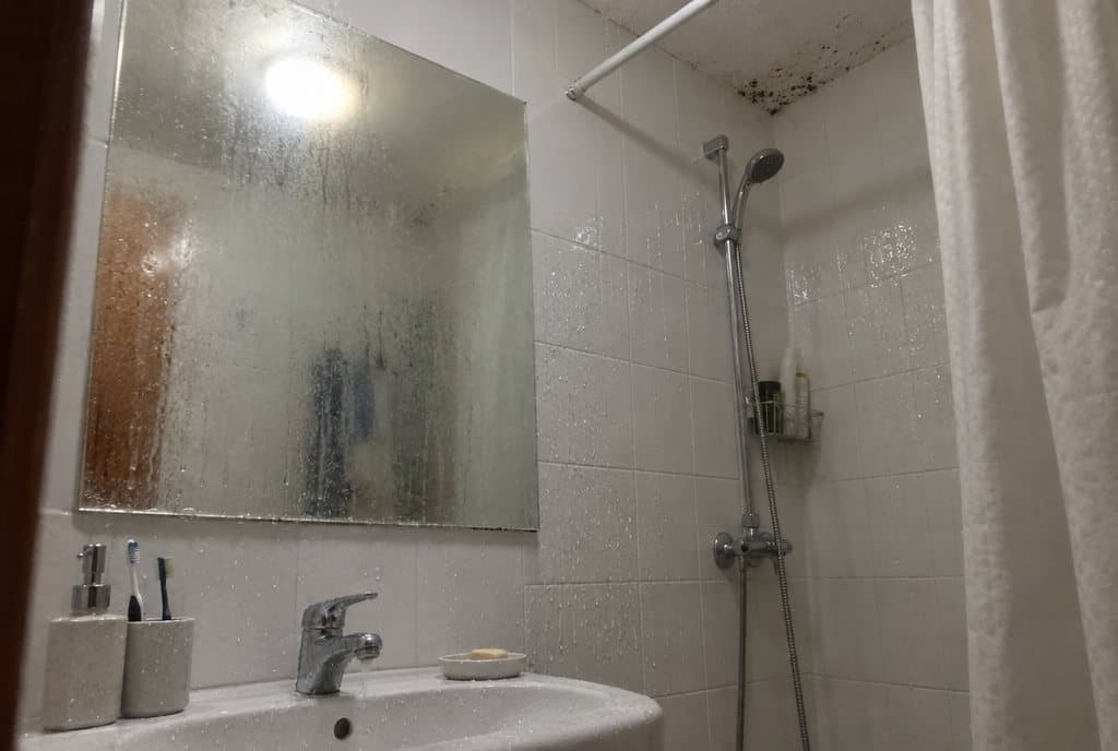 Baño interior después de una ducha: espejo empañado, gotas en el azulejo y manchas de moho en la esquina del techo