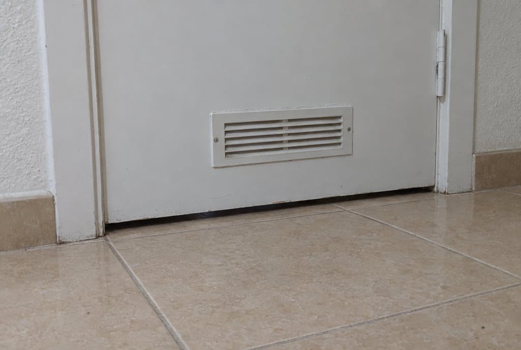 Parte baja de la puerta de un baño con holgura sobre el suelo y una rejilla de ventilación de plástico blanco