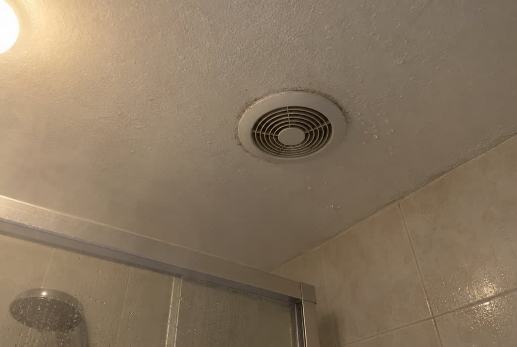 Extractor redondo blanco en el techo de un baño interior, con gotas de condensación alrededor y vapor sobre la mampara